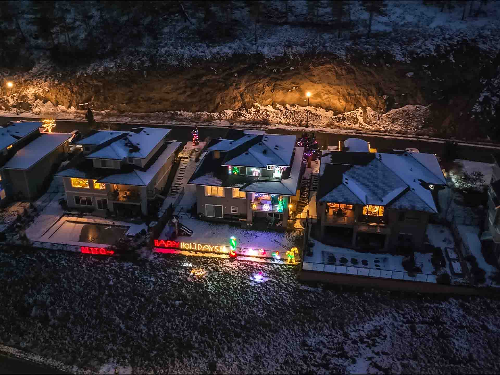 Happy Holidays lights on Wilden home with snowy homes and land around it, aerial perspective